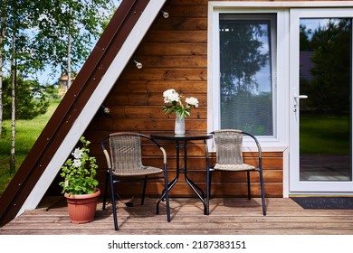 Cozy terrace with table and chairs near pot-plants. Wooden cottage house surrounded by greenery and trees in countryside on sunny summer day - Shutterstock ID 2187383151