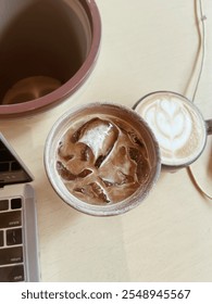 A cozy top-down view of a workspace featuring an iced coffee, a latte with latte art, and part of a laptop, set on a light wooden table, creating a relaxing coffee break vibe.
