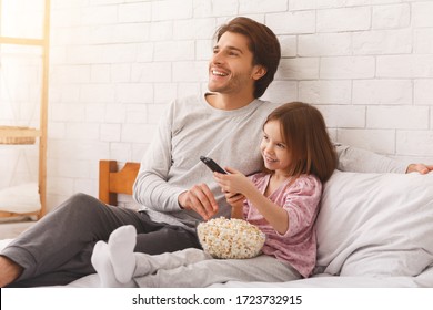 Cozy weekend at home. Cute little girl watching TV with her young dad, eating popcorn, copy space