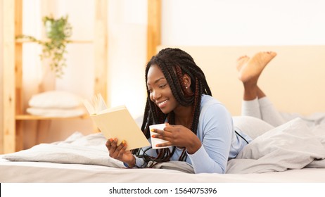 Cozy Weekend. Smiling African American Girl Reading Book And Drinking Coffee Lying In Bed At Home. Selective Focus, Panorama