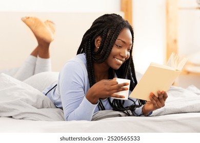 Cozy Weekend. Smiling African American Girl Reading Book And Drinking Coffee Lying In Bed At Home. Selective Focus, Panorama - Powered by Shutterstock