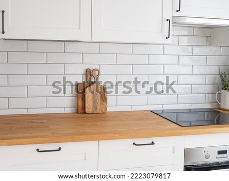 Cozy white kitchen with wooden countertop, flower and cutting boards in a minimalist interior with sunlight during the     day. Diagonal view.