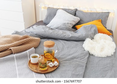 Cozy Winter Home Bedroom Decor: Glass Teapot And Two Cups Of Citrus Tea With Orange, Lemon And Mint On A Wooden Tray, Bed With Grey Linen, Blanket, Pillows, Plaid, Led Garland Light. Breakfast In Bed.