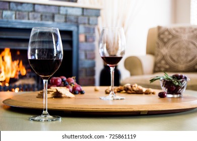 Cozy winter scene. Red wine and a snack platter with grapes, salted cracks, nuts and olives on a wooden board with a fire in a fireplace in the background. 