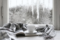 Cozy Winter Still Life: Cup Of Hot Coffee And Opened Book With Warm Plaid On Vintage Windowsill Against Frozen Big Icicles From Outside..