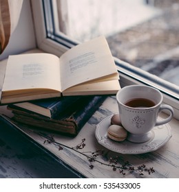 Cozy winter still life: cup of hot coffee and opened book on vintage windowsill against snow landscape from outside.