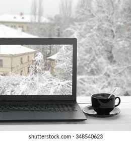 Cozy winter still life: laptop with outside view on screen and cup of hot coffee against snow landscape in window.