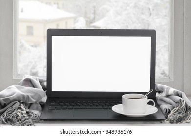 Cozy winter still life: laptop with blank screen, cup of coffee and warm plaid on windowsill against snow landscape from outside.
