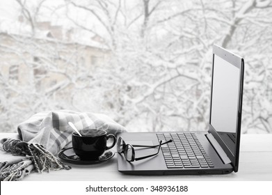 Cozy winter still life: laptop, glasses, cup of hot coffee and warm plaid on windowsill against snow landscape from outside.