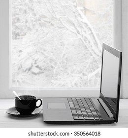 Cozy winter still life: laptop and cup of hot coffee on windowsill against snow landscape from outside.