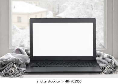 Cozy winter still life: laptop with blank screen and warm plaid on windowsill against snow landscape from outside.