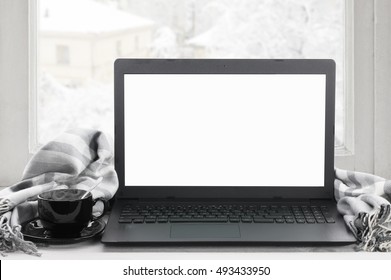 Cozy winter still life: laptop with blank screen, cup of hot coffee and warm plaid on windowsill against snow landscape from outside.