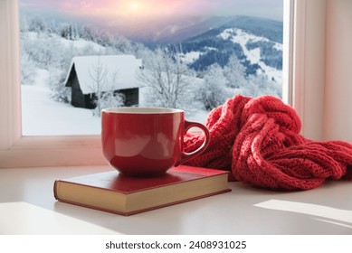 Cozy winter still life: mug of hot tea and opened book with warm plaid on modern windowsill against snow landscape from outside. Selective focus. High quality photo