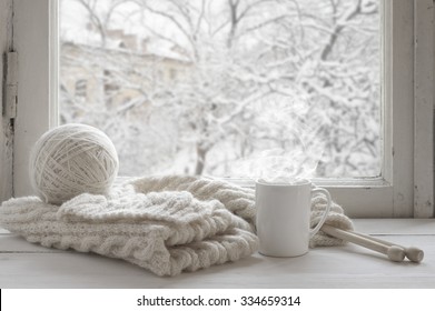 Cozy Winter Still Life: Mug Of Hot Tea And Warm Woolen Knitting On Vintage Windowsill Against Snow Landscape From Outside.