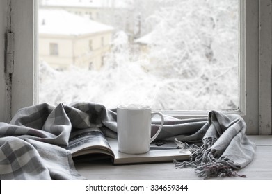 Cozy winter still life: mug of hot tea and opened book with warm plaid on vintage windowsill against snow landscape from outside.