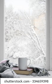 Cozy Winter Still Life: Mug Of Hot Tea And Book With Warm Plaid On Windowsill Against Snow Landscape From Outside.