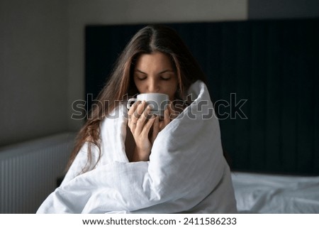 A cozy woman enjoys the warmth of a hot drink while wrapped in a blanket against the winter chill inside her bedroom.