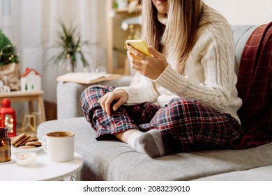 Cozy Woman In Knitted Winter Warm Socks, Sweater And Checkered Plaid With Phone, Drinking Hot Cocoa Or Coffee In Mug, During Resting On Couch At Home. Christmas Holidays With Candle, Decor And Cookies