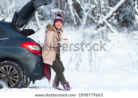 A cozy woman smiles, a female in warm winter clothes drinks a hot drink, tea or coffee, sits in the trunk of a car and smiles. Vacation, travel by car, snowy cold. Copy Space
