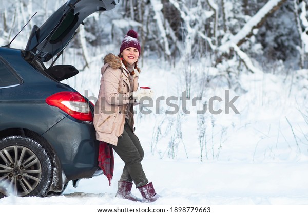A cozy\
woman smiles, a female in warm winter clothes drinks a hot drink,\
tea or coffee, sits in the trunk of a car and smiles. Vacation,\
travel by car, snowy cold. Copy\
Space