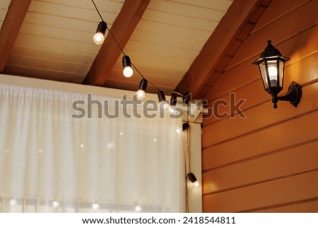 A cozy wooden patio is adorned with warmly lit string lights and a classic lantern. The front view against a white curtain backdrop. Concept for home decor or bilding cottages. 