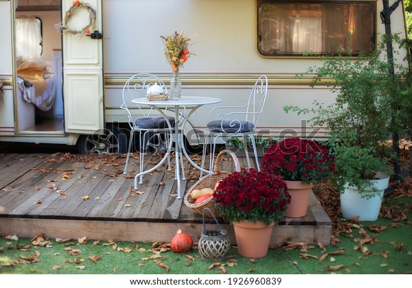 Cozy wooden RV house porch with garden furniture.\
Decor summer yard. Interior cozy patio with chrysanthemums in pots.\
Table and chairs with tea set  placed outside cozy retro caravan\
trailer in garden
