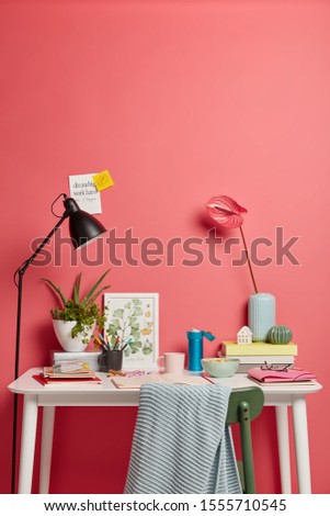 Cozy workplace with different things. Beautiful calla liles in vase, pile of books, opened diary with written notes, desk lamp, drawn picture, some food in bowl hot drink on white table over pink wall