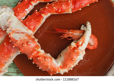 Crab Claw Grasping A Whole Cooked Shrimp On A Plate Accompanied By Additional Crab Legs To Crack Open For Their Meat