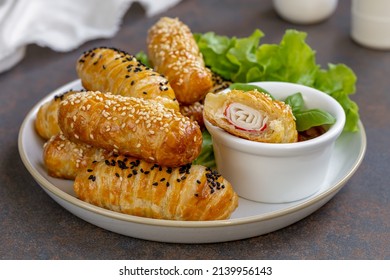 Crab Sticks in Puff Pastry. Baked puff pastry rolls or buns with filling served on a plate with green salad and tomato dipping sauce. Closeup with one broken open