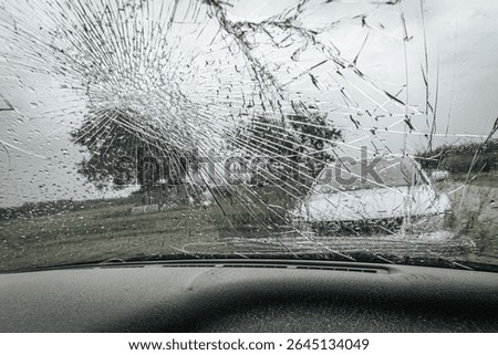 A cracked car windshield seen from the inside, with raindrops and a blurred outdoor scene including a white vehicle and green landscape in the background.