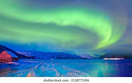 Cracks on the surface of the green ice next to white lone house (cabin) in winter - Landscape with house at night under green aurora sky - Northern lights (Aurora borealis) in the sky - Tromso, Norway - Shutterstock ID 2118771596