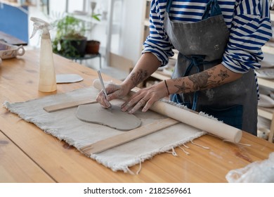 Craft artist making object out of clay in studio, cropped shot. Female ceramic artist with tattoo on hands use fettling knife to cut and trim clay, creating handmade kitchenware at pottery workshop - Shutterstock ID 2182569661