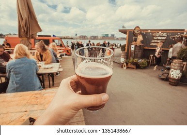 Craft Beer Glass In Drinker Hand On Street Food Market Of Copenhagen, Denmark. Leisure In Scandinavia With Drinks And Food Of Popular City Market.