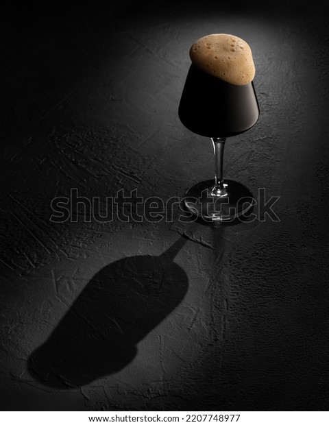 Craft Beer Stout Ale with foam in a\
cup. Dark Lager alcohol drink with rough black\
backdrop.