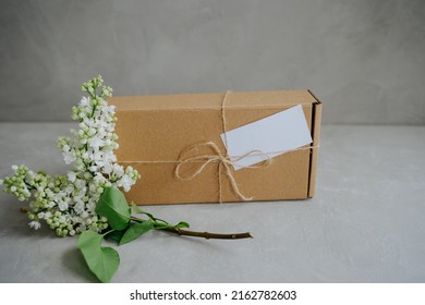 Craft Box Tied With Twine On A Bow. Delivery Of Goods To The Door. The Package. A Gift And A Branch Of White Lilac Flowers. 