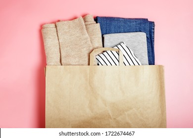 Craft brown paper bag with clothes (blue jeans, beige sweater, gray t-shirt, white shirt) on a pink background. Shopping concept, seasonal sale. Background for text, copy space.