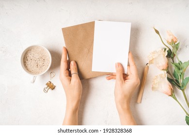 Craft Paper Envelope With White Blank Paper Note Mockup In Woman Hands, With Cup Of Coffee, Gold Binder Clips And Flowers On White Background. Flat Lay, Top View. Invitation, Package And Letter.