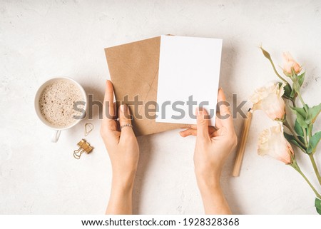 Craft paper envelope with white blank paper note mockup in woman hands, with cup of coffee, gold binder clips and flowers on white background. Flat lay, top view. Invitation, package and letter.