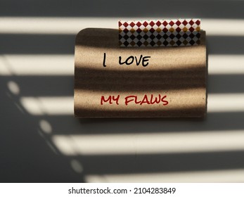 Craft paper stick on shadow background with handwritten text I LOVE MY FLAWS, self talk to remind to learn self acceptance, embeace imperfection, learn to love yourself.