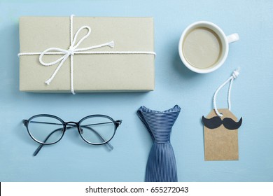 Craft Paper Wrapped Gift Box, Coffee, Glasses, Necktie And Craft Paper Card Tag With Mustache On Blue Background, Top View, Flat Layout As Hipster Style For Father's Day Concept