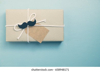 Craft Paper Wrapped Gift Box With Craft Paper Tag Card With Mustache On Blue Background, Top View, Flat Layout As Hipster Style For Father's Day Concept