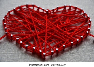 Craft String Art In Shape Of Heart. Red Woolen Heart, Symbol Of Love, Made Of Red Wool Yarn Threads Tangled Over Metal Nails On Canvas Background