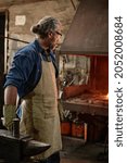 Craftsman in apron using work tools in his work while standing near the furnace in the workshop