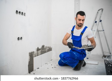 The craftsman attaches the tiles to the wall with cement. He is wearing a blue uniform and black gloves - Shutterstock ID 1837762966