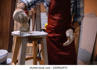 Craftsman, Carpenter Using A Paintbrush Painting To Wooden Bar Chair Handmade At Woodworking Workshop Or Carpentry Workplace. Selective Soft Focus On The Paintbrush.