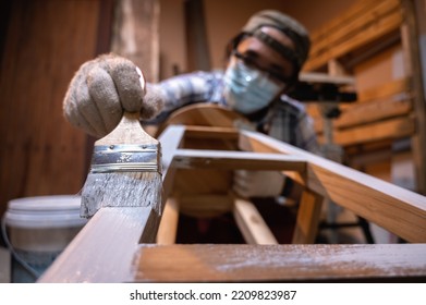 Craftsman, Carpenter Using A Paintbrush Painting To Wooden Bar Chair Handmade At Woodworking Workshop Or Carpentry Workplace. Selective Soft Focus On The Paintbrush.