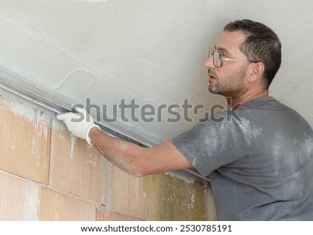 Craftsman checking the ceiling level and smooth surface with straight edge. Men in dirty gloves and gray shirt with dust and plaster on work clothing, construction process, back view