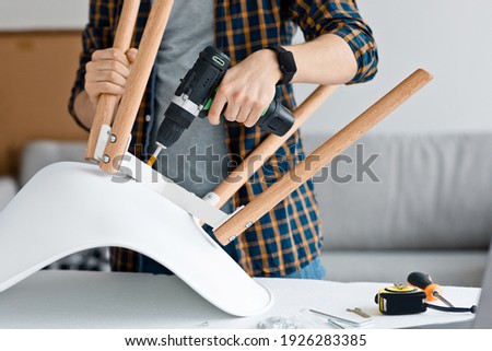 Craftsman collects and repairing with tools, assemble furniture with drill. Happy man screws details to chair with electric screwdriver in living room interior, close up, copy space, cropped