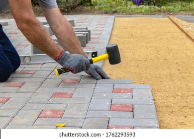 A craftsman in gloves with a rubber mallet lays concrete blocks on a gravelly sand base. Laying concrete blocks on the sidewalk.