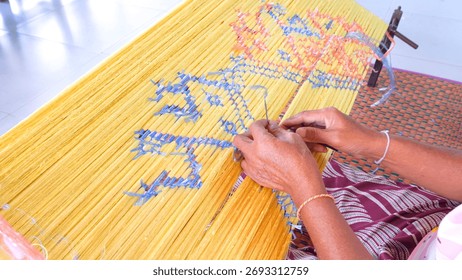 A craftsman tying plastic strings on silk threads to create traditional Thai Mudmee (ikat) patterns before dyeing. Handmade silk weaving process showing Thai culture, craftsmanship, and textile art.
